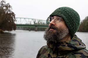 Bright photo of an older man looking wistful. There is a river and bridge behind him.